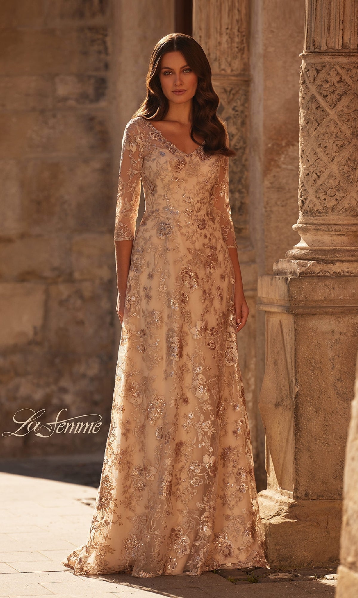 A woman stands outdoors in a champagne La Femme 30803 long floral-lace formal dress, featuring three-quarter sleeves and floral embroidery—ideal as a mother of the bride gown amid stone columns and textured walls.